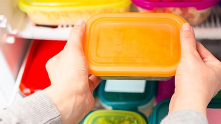 hands holding storage food container with open stocked fridge in background